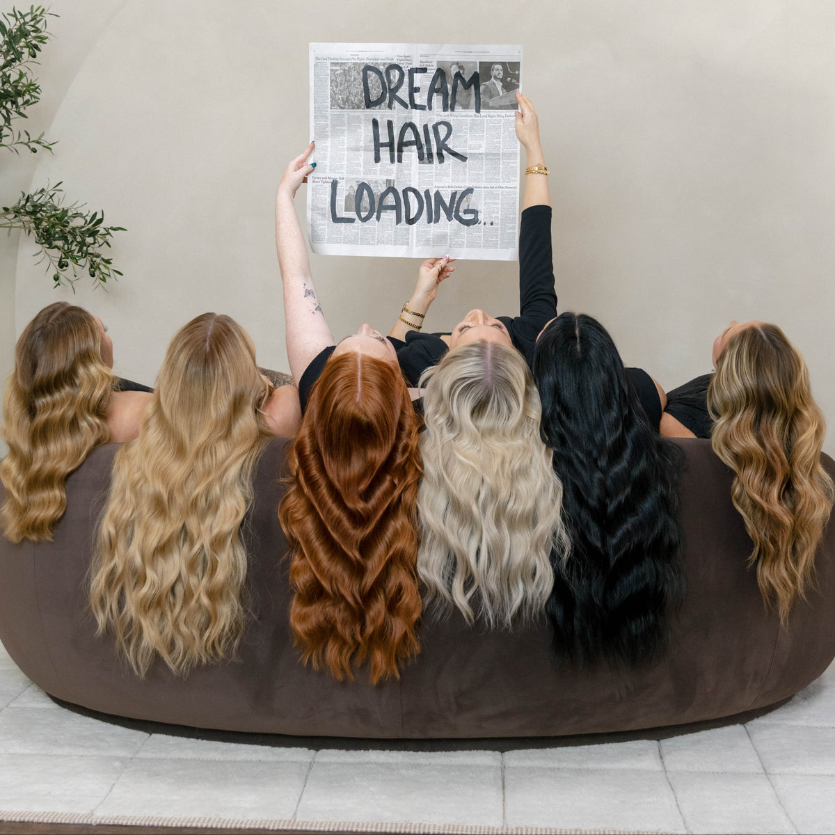 Five women with long wavy hair in different colors sit on a brown beanbag, holding up a sign that reads 'DREAM HAIR LOADING.